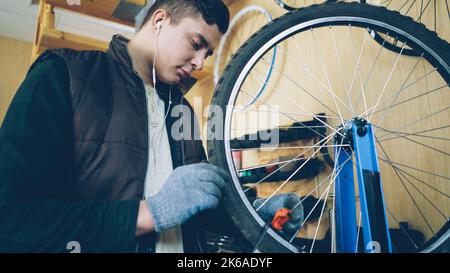 Concentrated mechanic is busy fixing spokes on bicycle wheel with special tools while servicing bike and listening to music through earphones. Maintenance and profession concept. Stock Photo