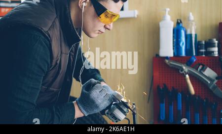 Side view of handsome young mechanic using electric circular saw in his busy workshop. Bright sparks, professional tools and equipment, safety uniform is visible. Stock Photo