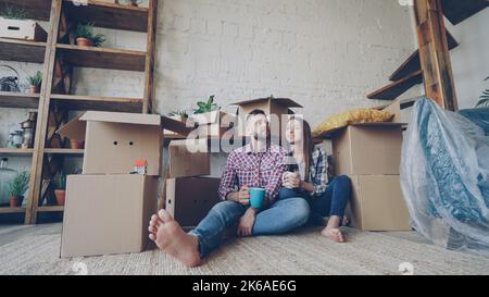 Adorable young couple is talking and holding mugs while sitting on floor of new flat after relocation. Numerous boxes, packed furniture and plants are visible. Stock Photo