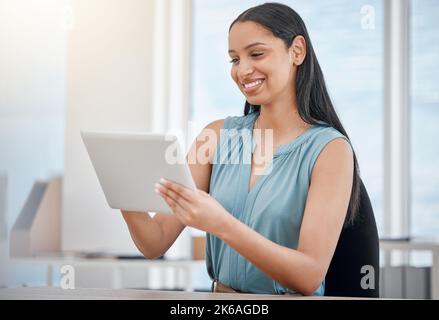 Young mixed race businesswoman smiling while working on a digital tablet in an office at work. One beautiful cheerful hispanic female boss using Stock Photo
