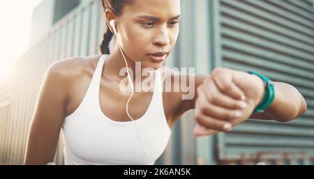 One active young hispanic woman wearing earphones and checking digital smartwatch while exercising in an urban setting outdoors. Female athlete Stock Photo