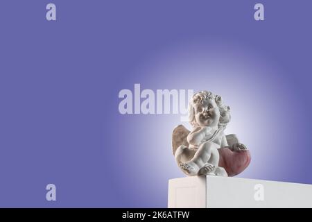 Figurine of an angel Cupid on the podium with a red heart on a very peri purple background . Valentine's Day. Stock Photo