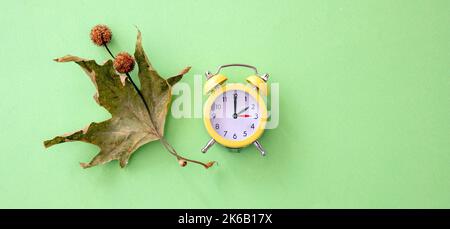 Fall Back, Daylight Saving Time. One hour back on yellow clock and autumn leaves on green background, top view Stock Photo