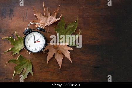 Fall Back, Daylight Saving Time. One hour back on yellow clock and autumn leaves on wooden background, top view Stock Photo