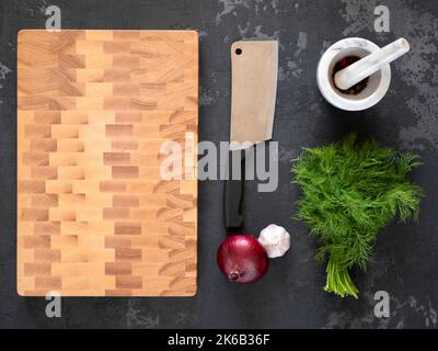Cutting board, knife, onion, garlic, dill and mortar with spices on dark background, flatlay, top view. Wooden block chopping board Stock Photo