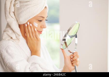 Woman with towel on head in bathroom looks and touches her face in the mirror and enjoys youth and hydration. Stock Photo