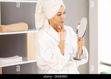 Woman with towel on head in bathroom looks and touches her face in the mirror and enjoys youth and hydration. Stock Photo