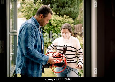 Man and woman talking to each other while holding electric car charger seen through doorway Stock Photo