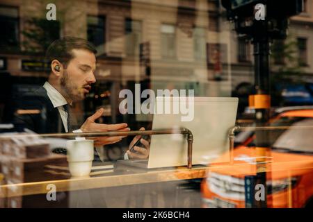 Young businessman gesturing while sitting with laptop in hotel seen through glass Stock Photo