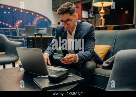 Businessman holding credit card while using laptop sitting in hotel lounge Stock Photo