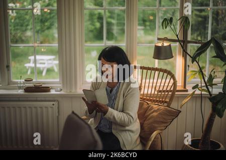 Mature woman using smart phone while sitting on chair near window at home Stock Photo