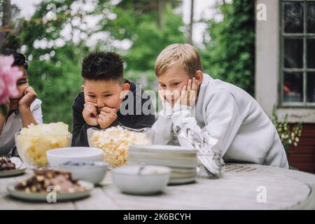 Curious boys leaning on elbows while looking at snacks bowl on table Stock Photo