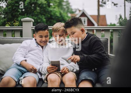 Happy male friends watching smart phone while sitting on couch Stock Photo