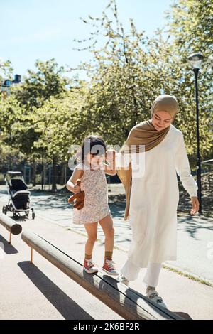 Mother holding hand of daughter while balancing on bench in park during sunny day Stock Photo