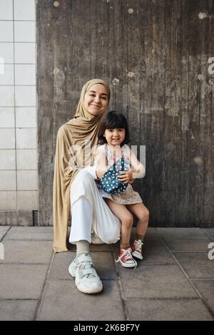 Smiling woman wearing hijab kneeling with daughter in front of wall Stock Photo