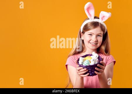 Studio Shot Of Girl Wearing Rabbit Ears And Holding Basket Of Easter Eggs Against Yellow Background Stock Photo