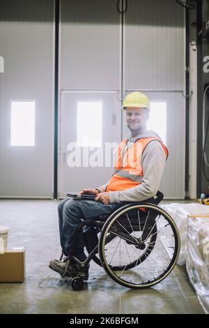 Portrait of smiling carpenter with disability holding digital tablet while sitting on wheelchair in warehouse Stock Photo
