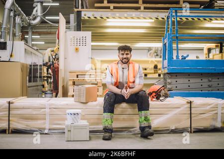 Portrait of smiling young blue-collar worker sitting by hardhat in warehouse Stock Photo