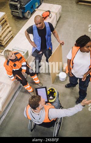 Blue-collar worker sitting on wheelchair instructing colleagues in warehouse Stock Photo