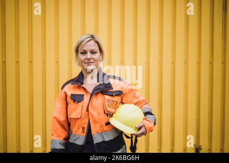 Portrait of mature female construction worker with hardhat against yellow metal wall Stock Photo