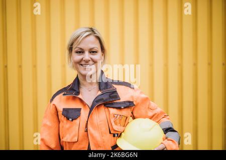 Portrait of happy female construction worker with hardhat against yellow metal wall Stock Photo
