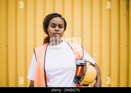 Portrait of confident female construction worker with hardhat against yellow metal wall Stock Photo