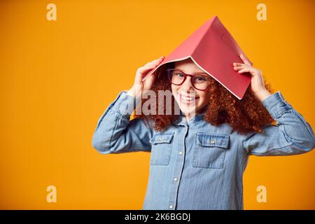 Studio Shot Of Girl Wearing Glasses With School Exercise Book On Head Against Yellow Background Stock Photo