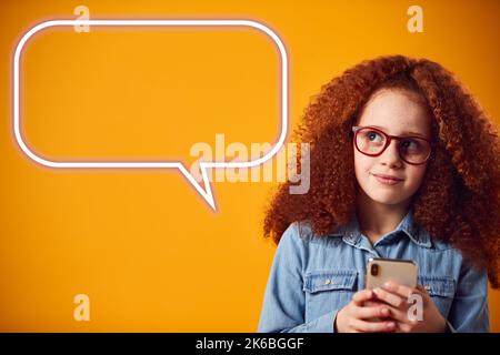 Studio Portrait Of Girl Using Mobile Phone With Speech Bubble Graphic Against Yellow Background Stock Photo