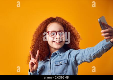 Studio Portrait Of Girl Wearing Glasses Posing For Selfie On Mobile Phone Against Yellow Background Stock Photo