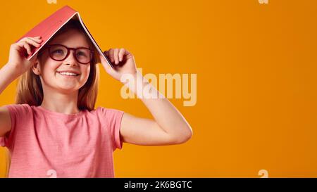 Studio Shot Of Girl Wearing Glasses With School Exercise Book On Head Against Yellow Background Stock Photo