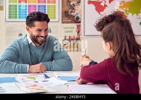 Smiling Male Elementary School Teacher Working At Desk In Classroom With Students Stock Photo