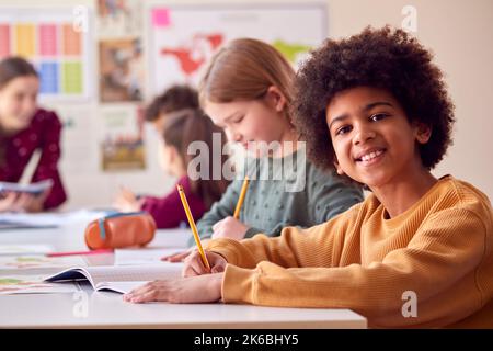 Portrait Of Smiling Male Elementary School Student Working At Desk In Classroom With Teacher Stock Photo