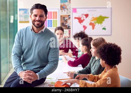 Portrait Of Smiling Male Elementary School Teacher Working At Desk In Classroom With Students Stock Photo