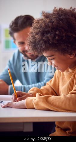 Teacher With Male Student In School Classroom Sitting At Desk Writing In Book Together Stock Photo