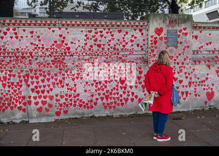 London, UK. 12th October, 2022. A woman in a red coat looks at the red painted hearts in memory of the thousands who have died from Covid-19 painted on The National Covid Memorial Wall next to the River Thames in London. The number of people dying within 28 days of testing postive for Covid-19 is sadly on the increase again. Credit: Maureen McLean/Alamy Live News Stock Photo