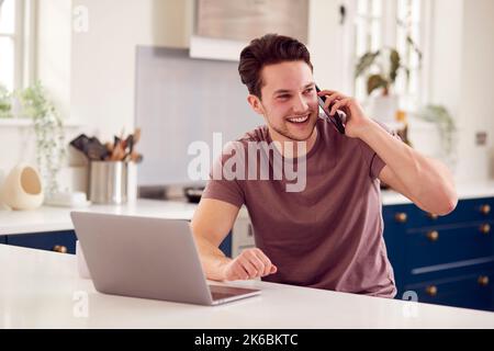 Transgender Man Using Mobile Phone Working From Home Using Laptop On Kitchen Counter Stock Photo