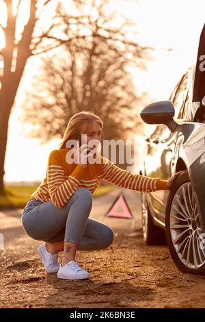 Woman Calling For Help On Phone After Car Breakdown On Country Road With Tyre Puncture Stock Photo