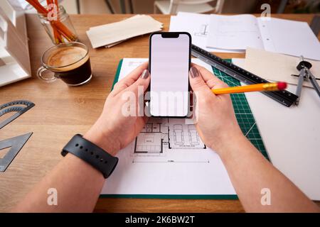 POV Shot Of Female Architect Working In Office On Plans For New Building Using Mobile Phone Stock Photo