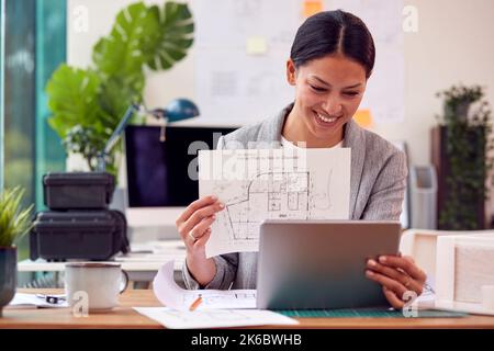 Female Architect In Office Sitting At Desk Showing Plans For New Building On Video Call Stock Photo