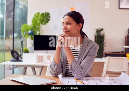 Thoughtful Female Architect In Office Sitting At Desk Working On Plans For New Building On Laptop Stock Photo