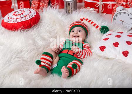 Newborn baby boy dressed in gnome costume lying on white fur carpet among christmas decorations. Christmas photo of infant in ctriped cap. New Year co Stock Photo