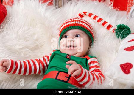 Newborn baby boy dressed in gnome costume lying on white fur carpet among christmas decorations. Christmas photo of infant in ctriped cap. New Year co Stock Photo