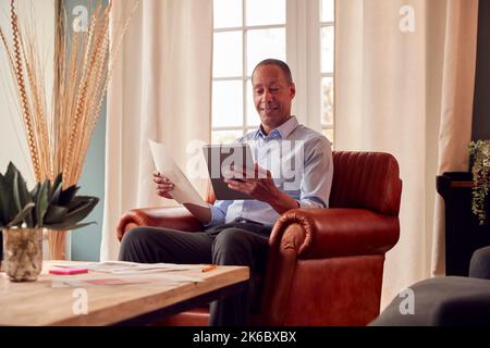 Mature Man Or Businessman Working From Home Sits In Armchair Reading Document Using Digital Tablet Stock Photo