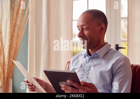 Mature Man Or Businessman Working From Home Sits In Armchair Reading Document Using Digital Tablet Stock Photo