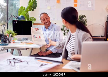 Male And Female Architects Working In Office At Desks Discussing Document On New Building Stock Photo