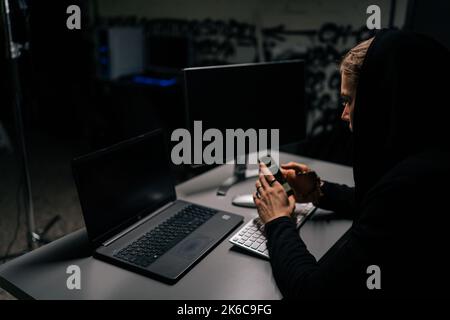 View from back to hidden hacker man wearing sweatshirt with hood engaged in hacking into security systems using mobile phone Stock Photo
