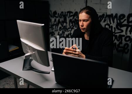 Portrait of young hacker man wearing long-sleeved shirt stealing information, preparing to break password sitting at desk in dark room. Stock Photo