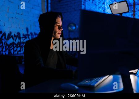 Side view of dangerous hacker male breaking into corporate data servers using laptop and computers sitting at desk in dark room with blue neon lights. Stock Photo