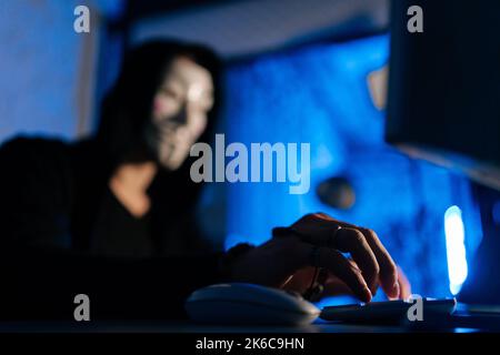 Selective focus of unrecognizable hooded hacker man wearing anonymous mask using laptop and desktop computer to hack network system. Stock Photo