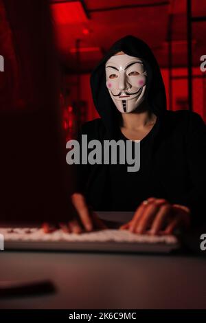 Close-up vertical shot of wanted hacker man wearing anonymous mask and sweatshirt with hood engaged in hacking into security systems, Stock Photo